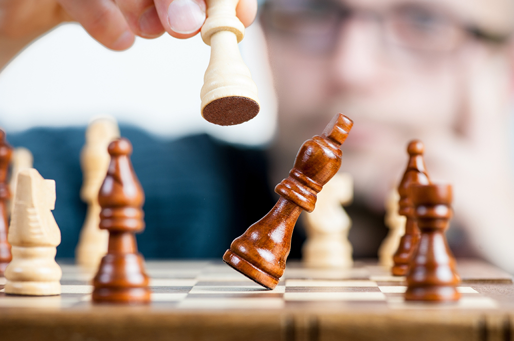 Ein Schachspiel, mit einer Hand, die eine Figur in die Höhe hält, während der König fällt. Das steht sinnbildlich für die Konkurrenzanalyse im Marketing.