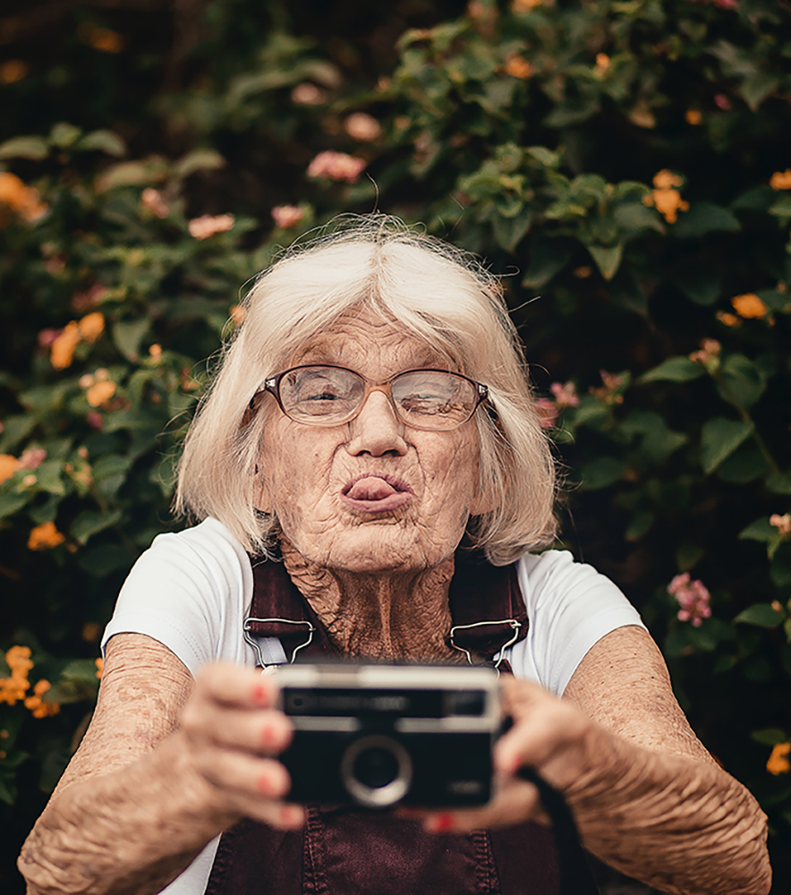 Eine ältere Dame mit Brille macht ein Selbstportrait mit einer Kamera und steckt dabei die Zunge heraus.