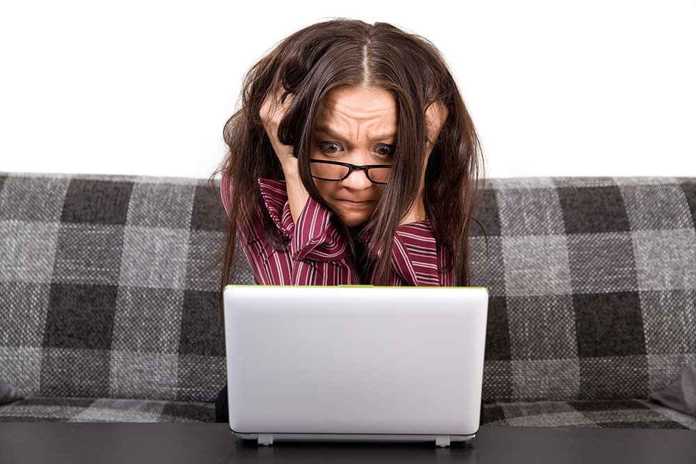 Verzweifelt aussehende Frau sitzt auf ihrer karierten Couch vor ihrem Laptop und fasst sich in die Haare. Ihre Brille hängt an der Nasenspitze, sie starrt in den PC mit faltiger Stirn.